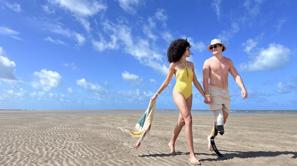 Casal caminha na areia sob céu azul de praia em Japaratinga.
