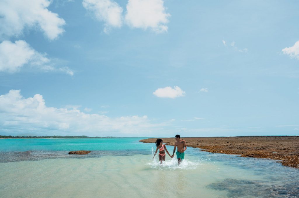 Casal caminha nas águas cristalinas entre recifes de corais das piscinas naturais de Japaratinga.