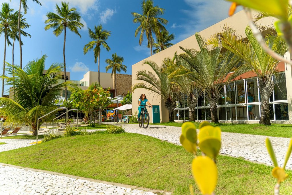 Trabalhadora de Experiência e Wellness do Japaratinga Lounge Resort anda de bicicleta em frente à Academia do resort All Inclusive.