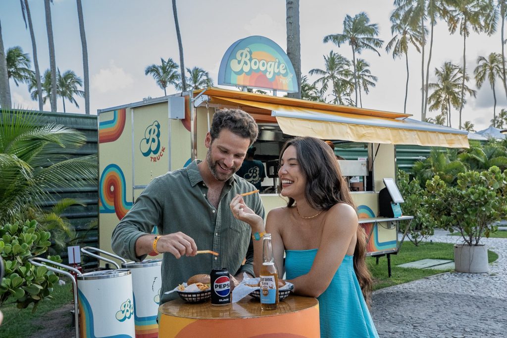 Casal desfrutando combo completo de hamburguers, batatas fritas e bebidas no Boogie Truck, ponto de consumo do Japaratinga Lounge Resort.