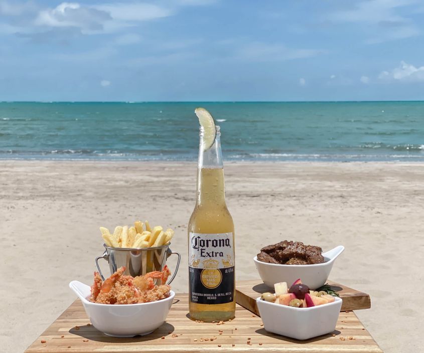 Petiscos e cerveja Corona servidos à beira-mar, em um ambiente paradisíaco de praia.