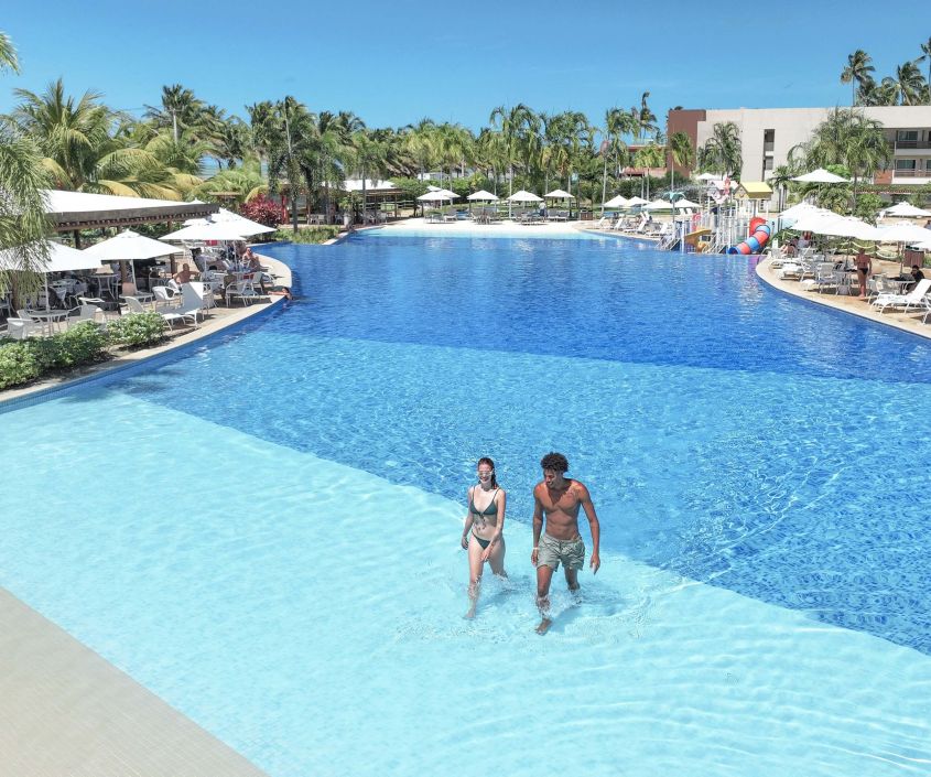 Casal caminhando na piscina da nova categoria Garden no Japaratinga Lounge Resort, cercado por uma área relaxante com espreguiçadeiras e guarda-sóis.