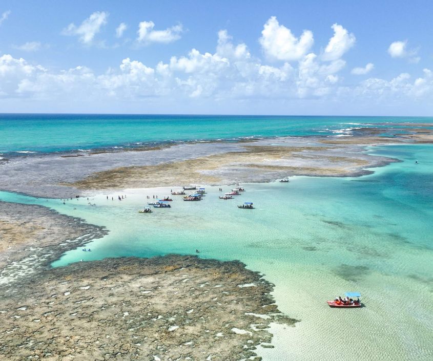Vista aérea das piscinas naturais de Japaratinga, com águas cristalinas e jangadas levando turistas para explorar a beleza do local.