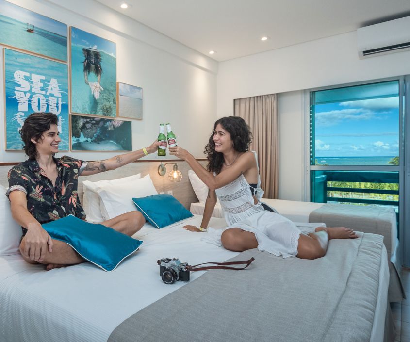 Um casal está brindando em cima da cama de um dos apartamentos do Japaratinga Lounge Resort. É possível ver parte do quarto e sua decoração jovial, também parte de sua varanda que oferece uma vista para a beira-mar de japaratinga.