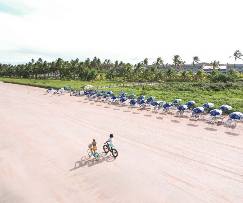 Ciclistas na praia de Japaratinga ao lado de uma fileira de cadeiras e guarda-sóis do Japaratinga Lounge Resort.