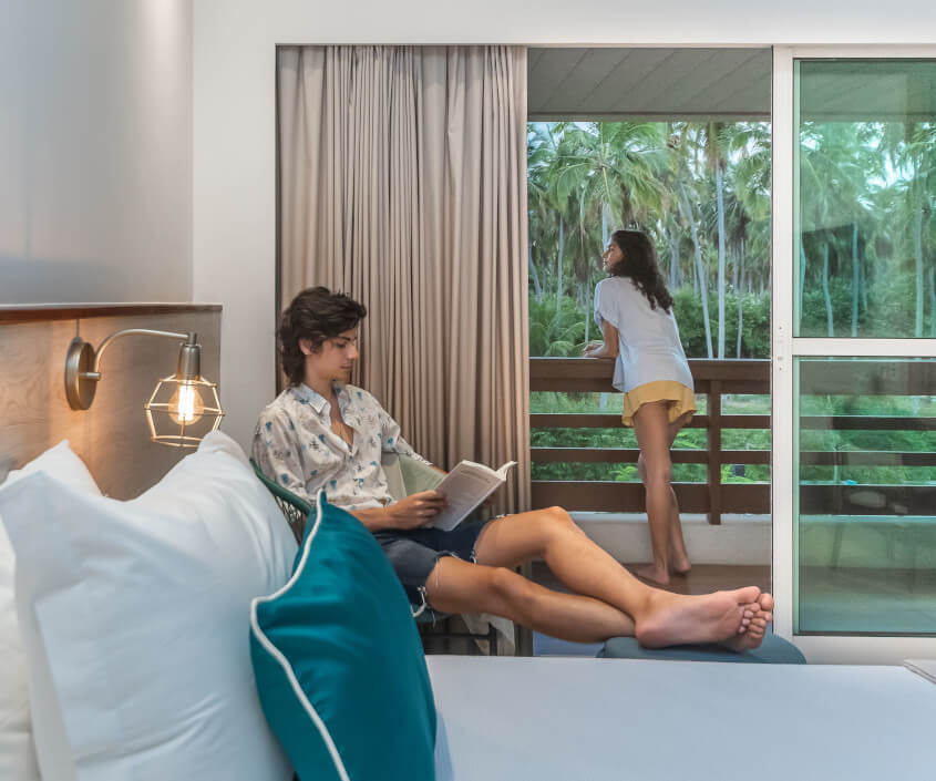 Homem sentado na cadeira lendo um livro, com pés apoiados no puf e mulher na varanda contemplando a vista.