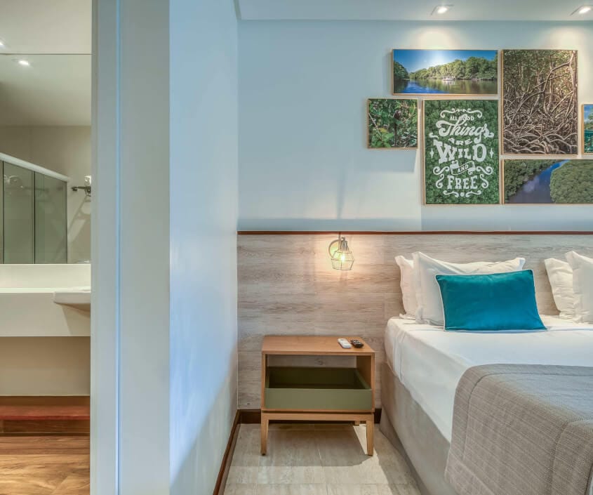 Entrada do banheiro, mesa de cabeceira com luminária e controles, e cama com quadros decorativos acima de temática mangue.