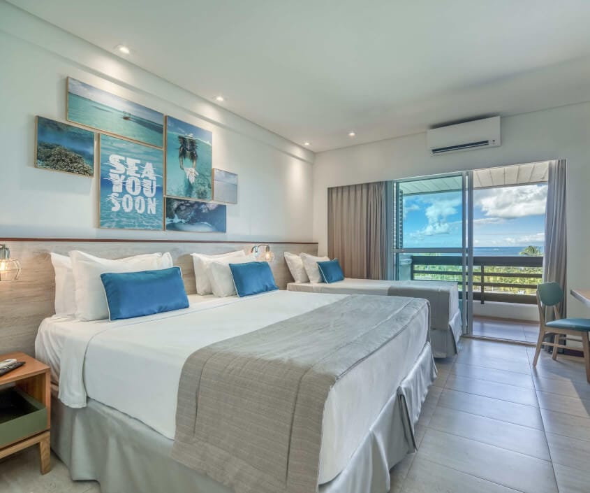 Apartamento Standard, com uma cama de casal e uma de solteiro, mesa para apoio, mesa de cabeceira, varanda com vista mar e piscina e decoração em tons de azul.