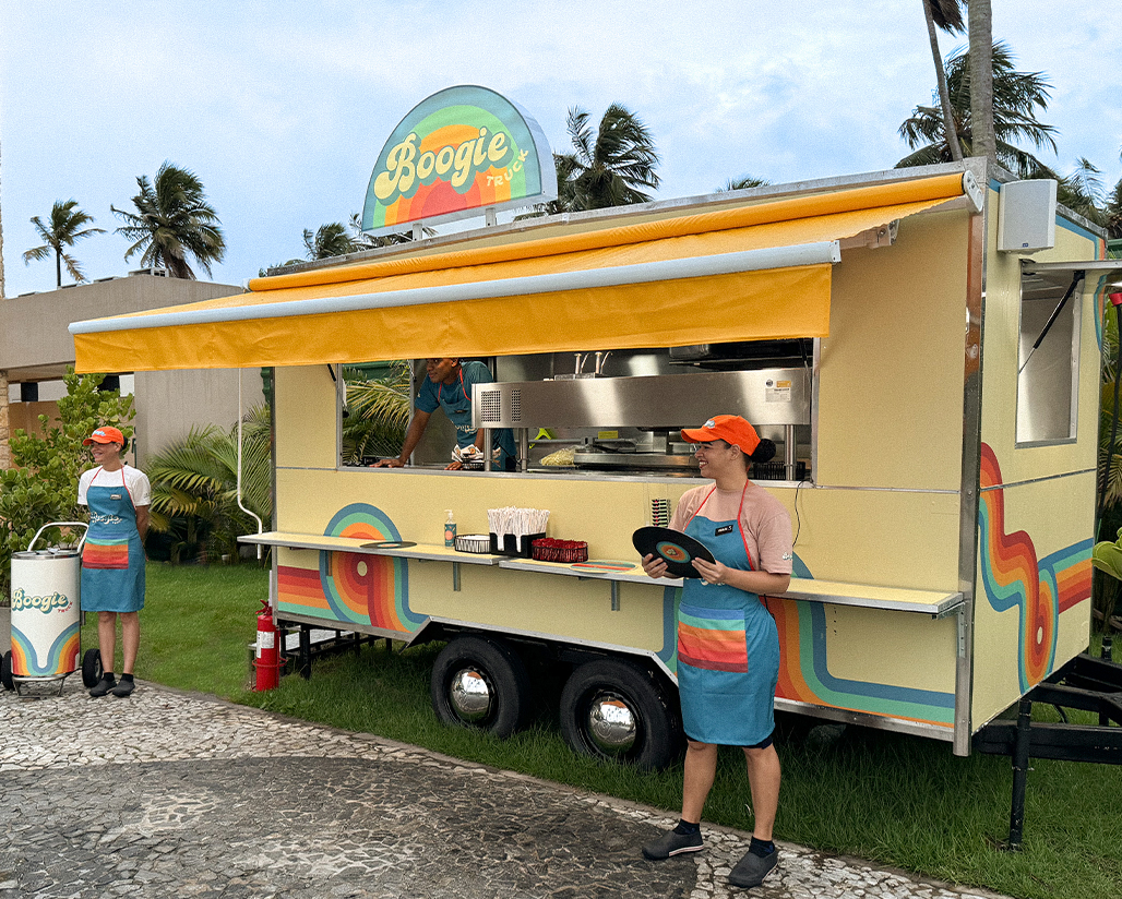  Food truck amarelo com um design colorido e retrô, chamado Boogie Truck. Com um funcionário sorri da janela de serviço e duas funcionárias na parte da frente do food truck, com palmeiras e um céu azul com algumas nuvens.
