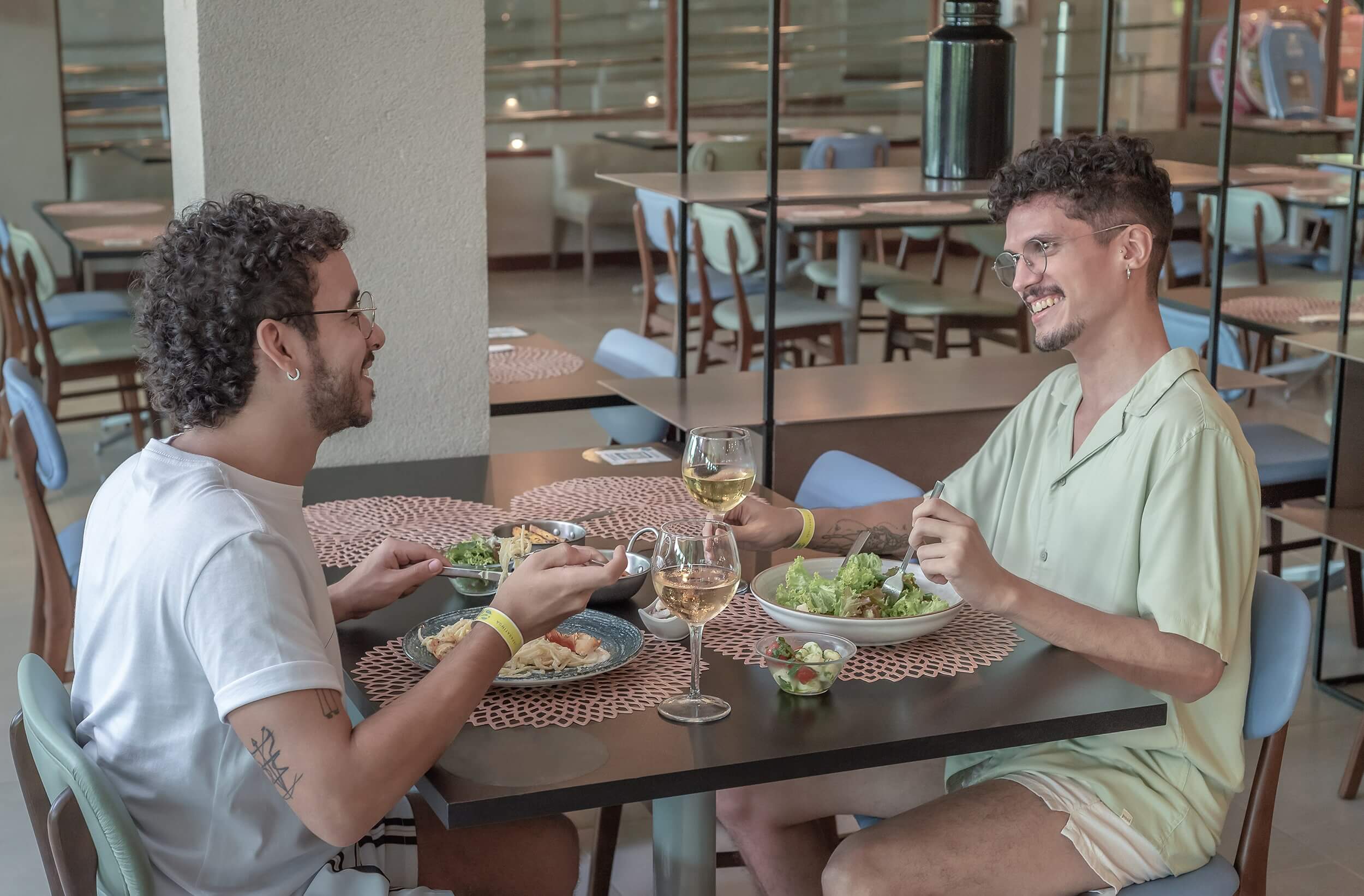 Dois homens em um restaurante, desfrutando de uma refeição. Ambos de cabelos cacheados e um de camiseta branca, o outro de camisa clara. É um ambiente moderno, e há taças de vinho na mesa.