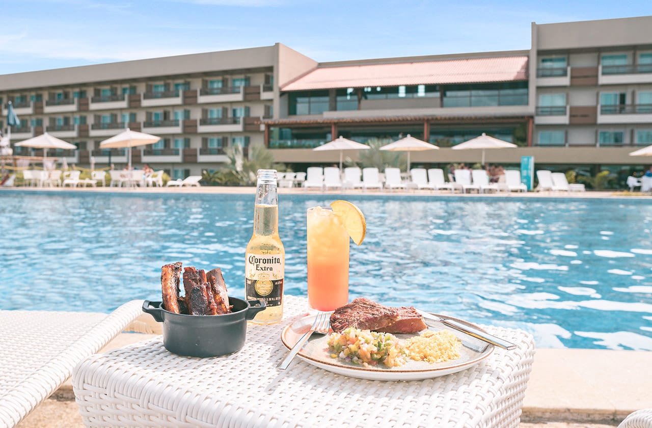 Uma mesa à beira de uma piscina  tem uma refeição que inclui costelas grelhadas em uma panela de ferro, uma garrafa de cerveja Coronita e um prato com um pedaço de carne acompanhado de arroz e salada. Ao lado, um coquetel laranja com uma fatia de limão.