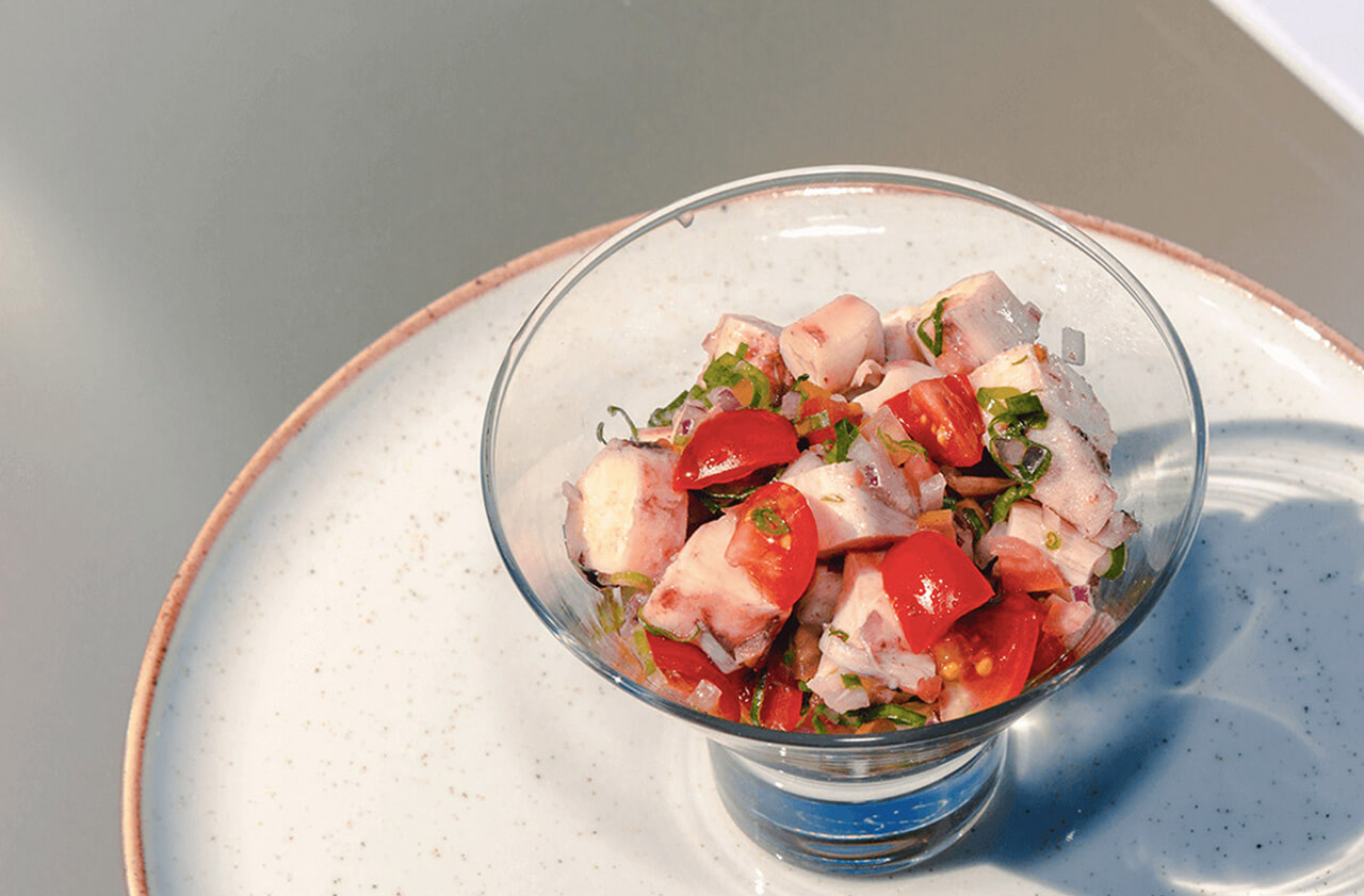 A imagem mostra uma taça de vidro contendo uma salada composta por pedaços de peixe, tomates-cereja cortados ao meio e cebolinhas. A taça está posicionada em um prato de cor clara. O plano de fundo da imagem tem uma cor neutra.