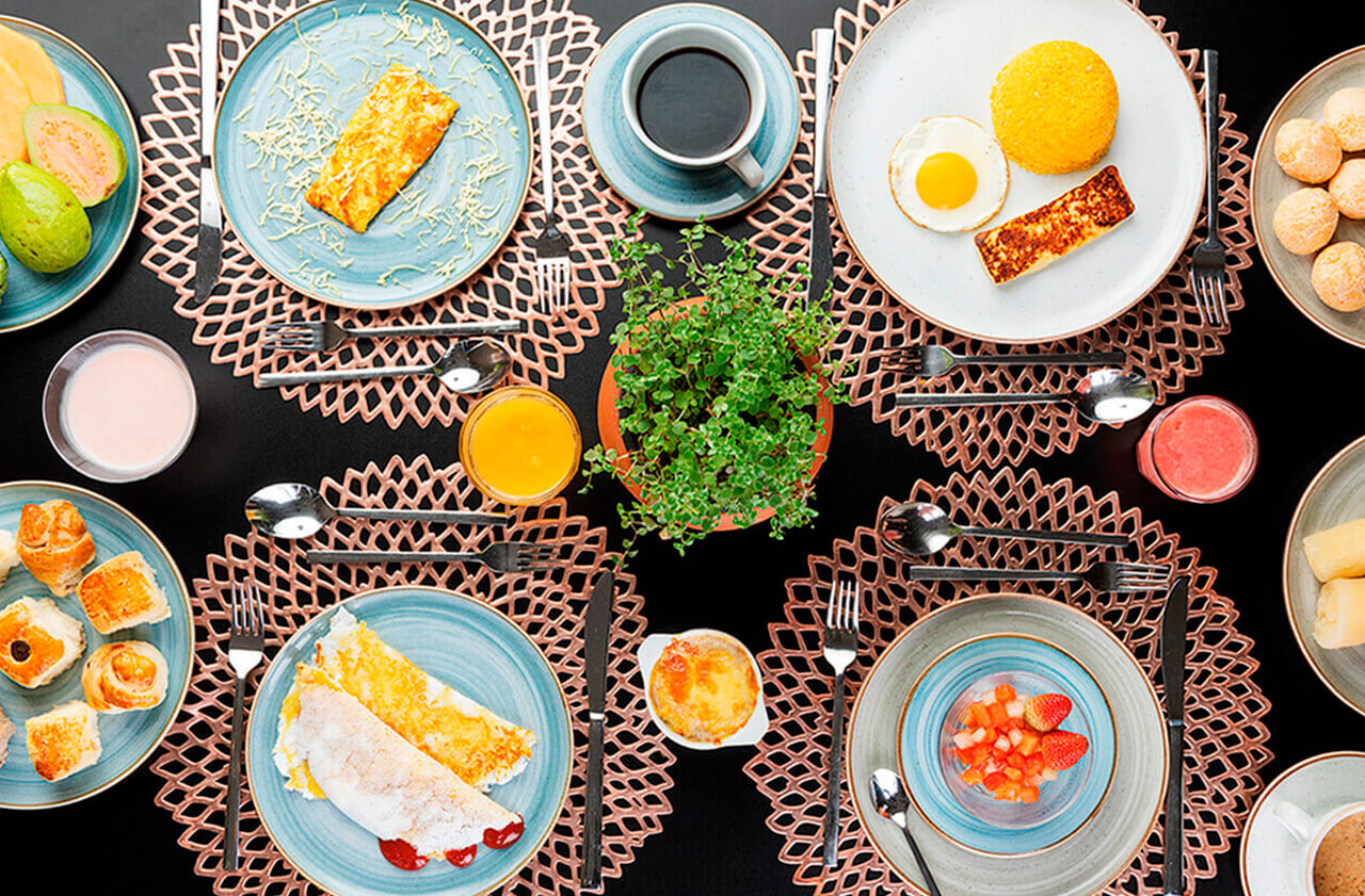 Mesa posta com omelete, ovo frito, sucos, café, panqueca com geleia e frutas. Design geométrico e detalhes requintados. Há também garfos, facas, colheres e uma xícara com café.