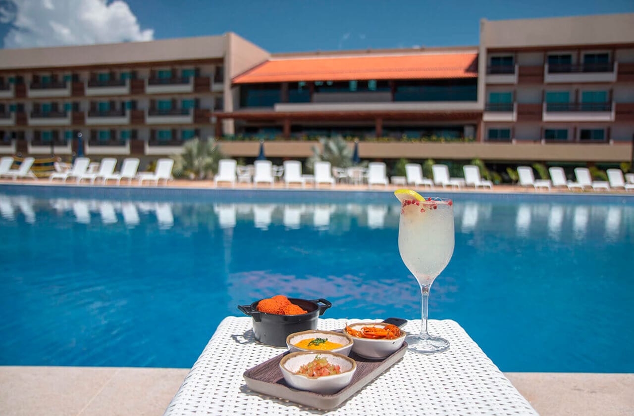 À beira de uma piscina, há uma mesa com uma bebida refrescante com uma fatia de limão e especiarias. Ao lado, pratos gourmet incluem croquetes, molhos variados e outros petiscos. Ao fundo, vê-se um prédio moderno e espreguiçadeiras alinhadas.