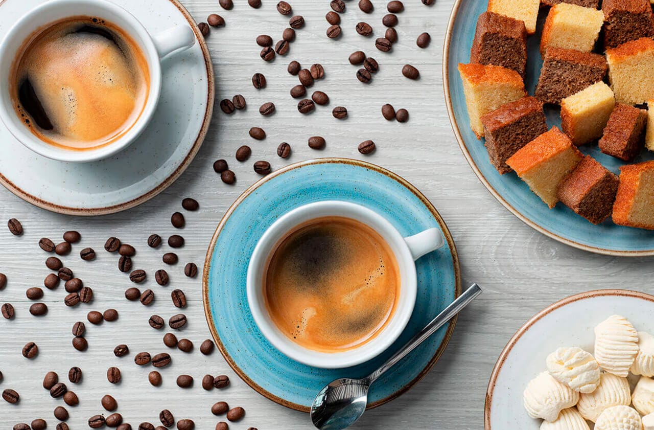 Na imagem há duas xícaras de café expresso, grãos de café espalhados ao fundo, há também um prato com pedaços de bolo alternados e outro com uma porção de bolo de goma.