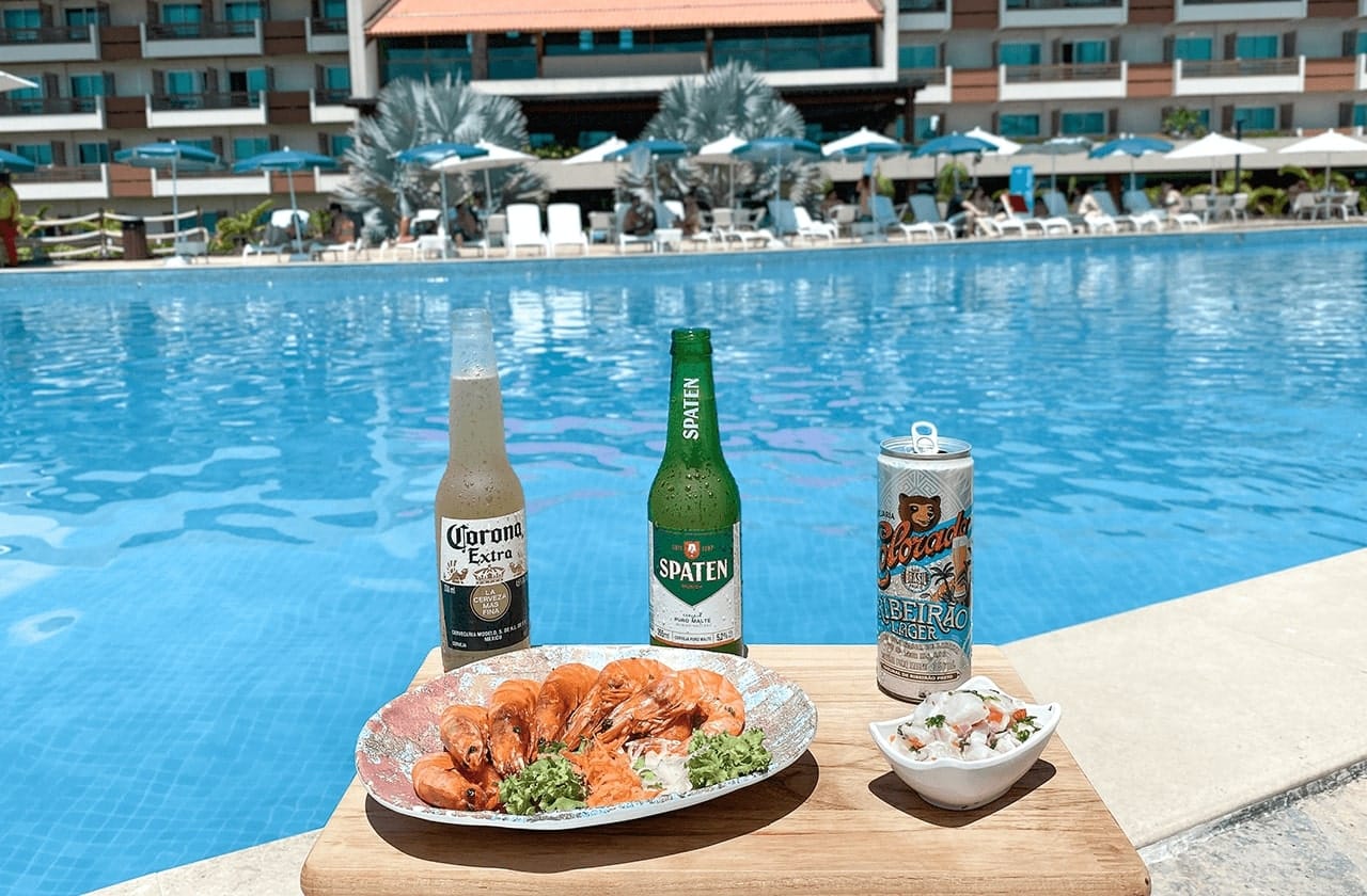 Seleção de bebidas e petiscos na beira de uma piscina. Há uma tábua sobre o qual estão uma garrafa de cerveja Corona, uma de cerveja Spaten e uma lata de Ribeirão Lager. Ao lado, há um prato com camarões cozidos e uma  tigela de salada de frutos do mar. Desfocado ao fundo há uma piscina azul.