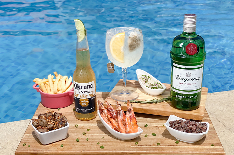 Bandeja de madeira com Gin Tônica, cerveja, garrafa de Gin Tanqueray, camarões, batatas fritas e porções de petiscos, servida na borda de uma piscina azul.