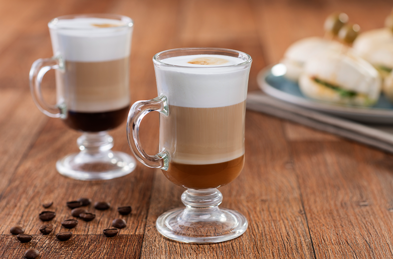 Duas canecas de vidro com café cremoso e em camadas, sobre uma mesa de madeira. Grãos de café e pequenos sanduíches são visíveis no fundo.