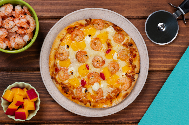 Uma pizza gourmet de camarão e queijo do reino em um prato, com uma tigela de camarões extras, mais queijos picados ao lado e o equipamento pra cortar.