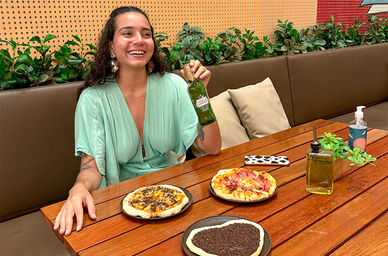 Uma mulher sorridente, sentada em um banco de couro com uma parede com mural colorido. A mesa tem três mini pizzas (doce e salgadas) e azeites.