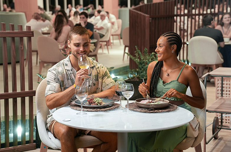 Um casal sorridente desfruta de um jantar romântico em um restaurante moderno de resort com iluminação suave. O homem segura uma taça de vinho branco.