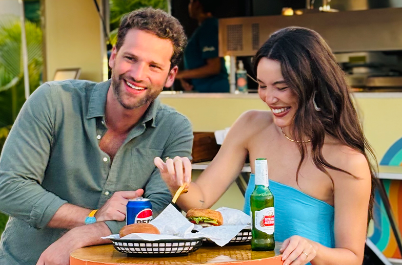 Um casal sorridente desfruta de hambúrgueres e cerveja em frente a um food truck de estilo retrô (Boogie Truck). O casal está em um ambiente tropical ensolarado com coqueiros e céu azul.