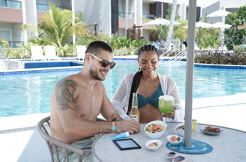 Um casal jovem e sorridente relaxa em uma mesa à beira de uma piscina de resort. Há drinks, cerveja, um kindle, e pequenas tigelas com petiscos e frutas na mesa.