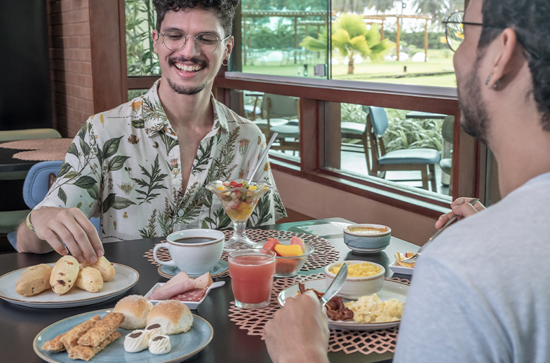 Dois jovens desfrutam do café da manhã. A mesa está cheia de pratos com pães de queijo, frutas, ovos e café. A janela de trás mostra a área verde do resort.
