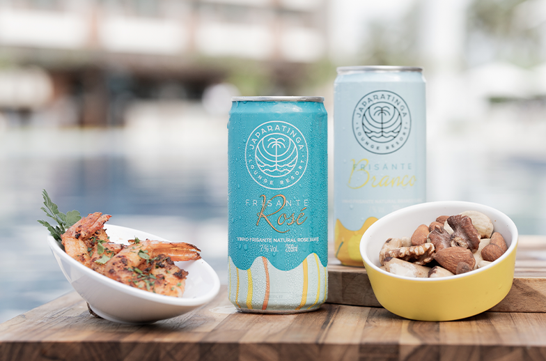 Duas latas de Frisante (Rosé e Branco) com a marca Japaratinga Lounge Resort sobre uma tábua de madeira. Ao lado, pratos com camarões grelhados e mix de castanhas à beira da piscina.