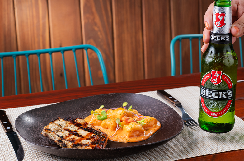 Prato gourmet com frango grelhado e gnocchi em molho. Uma mão segura uma garrafa de cerveja Beck's ao lado. As paredes e mesa do local são de madeira e as cadeiras são azuis.