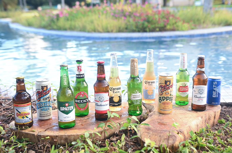 Uma grande variedade de cervejas em garrafas e latas (incluindo Stella Artois, Beck's, Spaten e Corona) dispostas sobre madeira na beira de uma piscina de resort.
