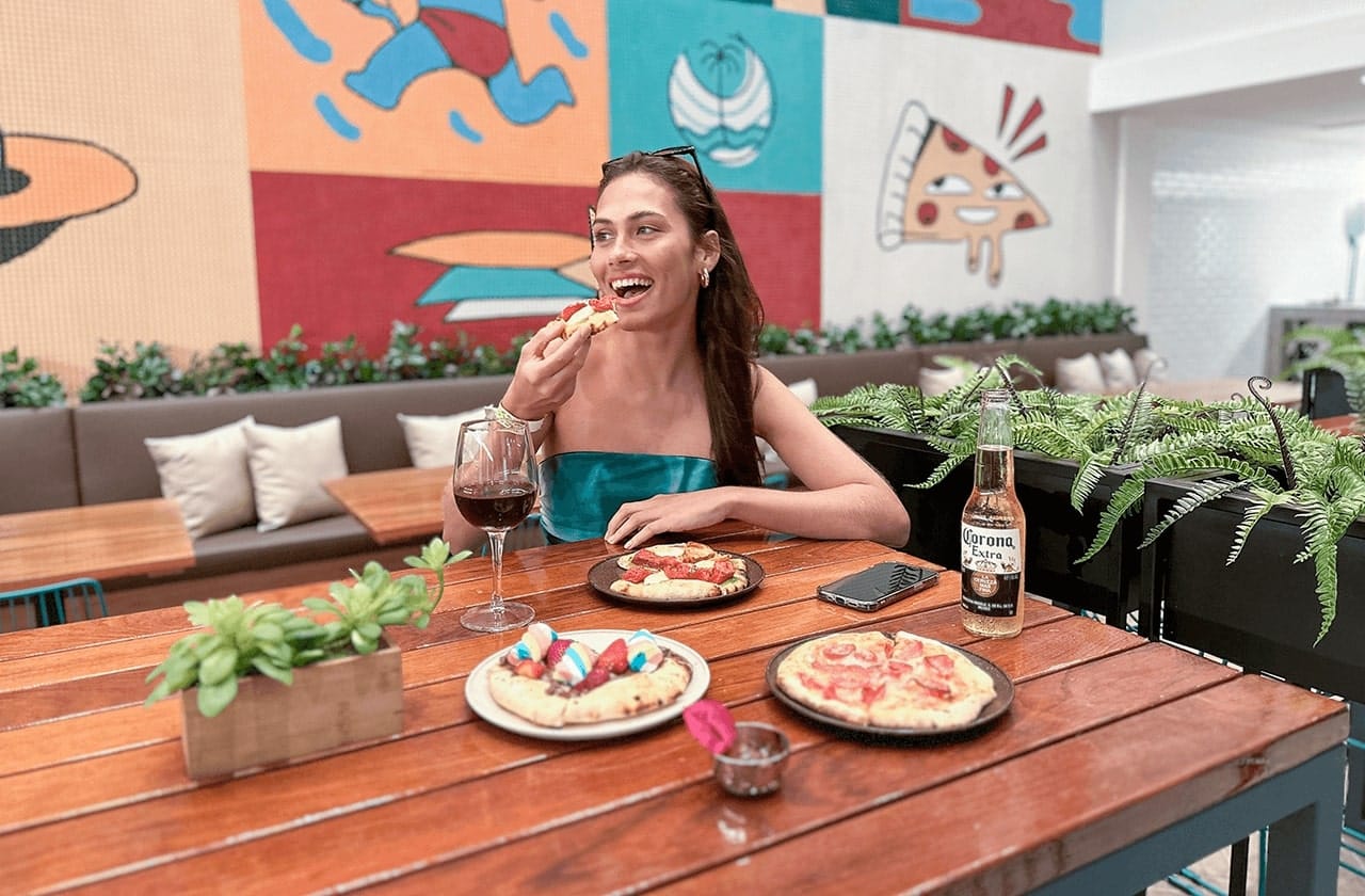 A imagem retrata uma mulher jovem sorridente, sentada à mesa em um restaurante descontraído e colorido. Ela está comendo pizza e tem à sua frente um copo de vinho tinto e uma garrafa de cerveja Corona e pizzas. Ao fundo, há um  grande mural colorido que apresenta diversas imagens lúdicas, como figuras humanas e um pedaço de pizza.