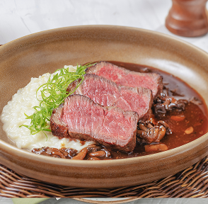 Prato com cortes de carne, purê e molho, servido em uma tigela de cerâmica e talheres ao lado sobre um jogo americano artesanal. Uma mão segura um copo com um coquetel ao lado.