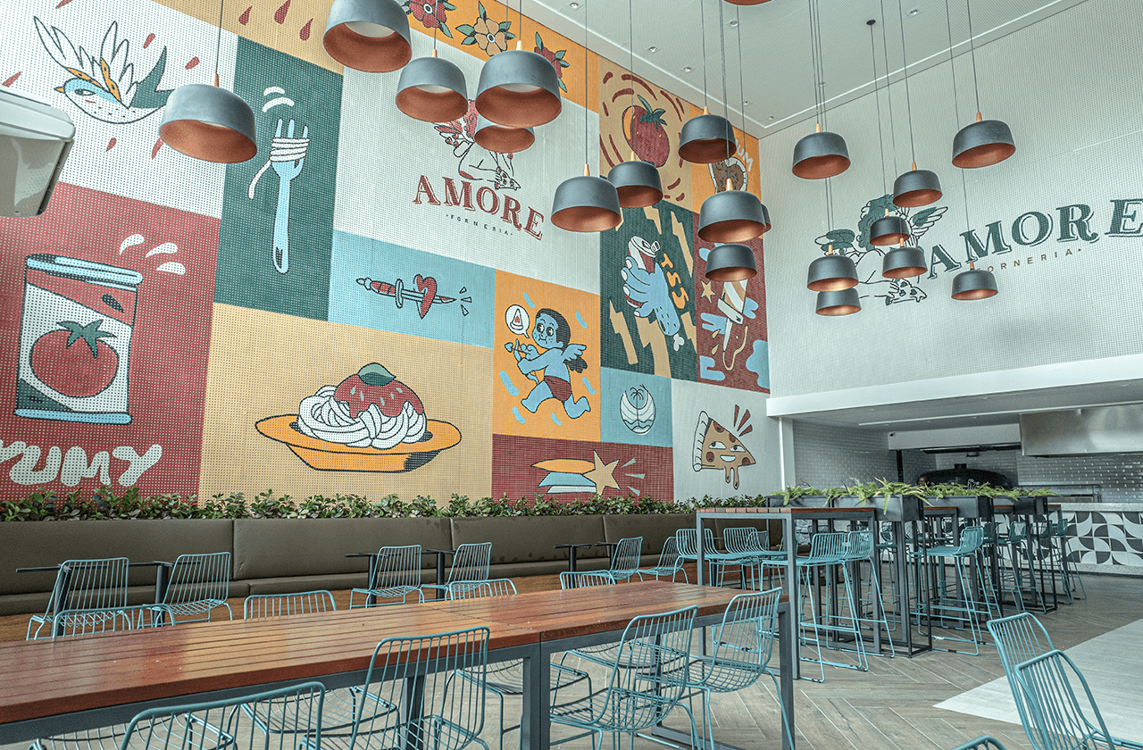 A imagem mostra a pizzaria Amore um ambiente jovial e moderno. Possui um grande mural colorido, com elementos gráficos estilizados que incluem alimentos, a frase "Amore Pizzeria", e ilustrações. Há mesas com cadeiras de metal azul claro, sob uma série de luminárias pendentes de design industrial em cobre.