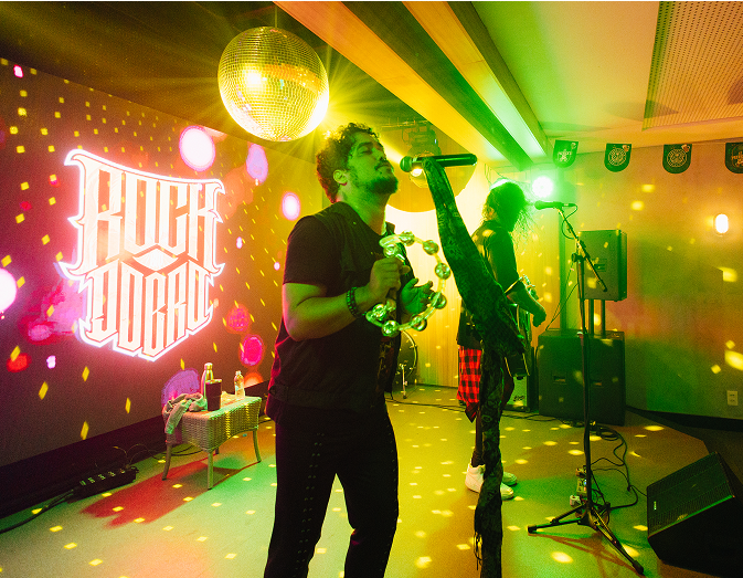 Atrações imperdíveis pra curtir a noite no melhor resort do Brasil Banda tocando com letreiro ao fundo e uma pequena mesa com bebidas em cima. O cantor está com uma pandeirola e o outro membro toca um violão. Luzes coloridas, e há globo de discoteca pendurado no teto no Pub Overdrive no Japaratinga Lounge Resort.