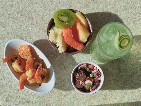 Vista de cima de uma mesa com tigelas de camarão grelhado, salada de frutas e salada de polvo, ao lado de dois coquetéis verdes.