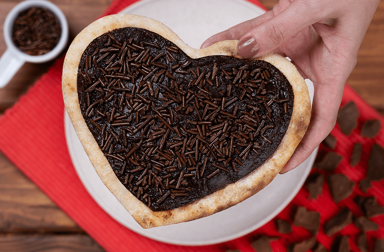 Há uma mão segurando uma pizza formato de coração. ela tem uma crosta dourada e é recheada com de chocolate, coberto com granulado de chocolate. Ela está sendo levantada de um prato branco, que está sobre um guardanapo ou pano de prato vermelho com textura. Ao fundo, pode-se ver uma mesa de madeira e uma tigela com mais granulado