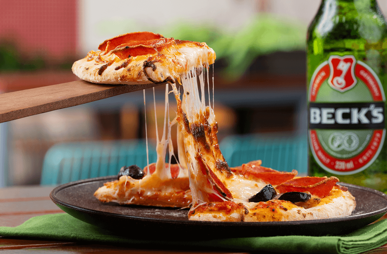 A imagem mostra uma fatia de pizza sendo levantada com uma pá de pizza, com o queijo esticando-se em fios longos e derretidos. Ela tem fatias de pepperoni e azeitonas pretas. Ela está em prato marrom. Há uma cerveja Beck's ao lado.