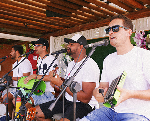 Grupo de quatro músicos brasileiros de samba e pagode em uma apresentação ao vivo no Bar Verano em Japaratinga Lounge Resort, todos vestindo camisetas brancas e cantando em microfones individuais enquanto tocam instrumentos de percussão e cordas, como tan-tan verde e cavaquinho.