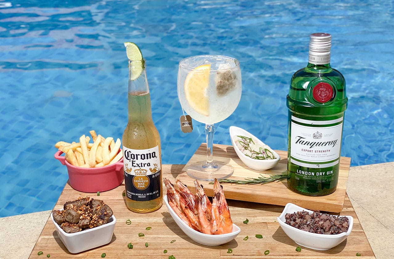 Pequena mesa na borda da piscina com cerveja Corona, porção de caudas de lagosta e drink Sex On The Beach. Canga, livro e óculos de sol ao redor.