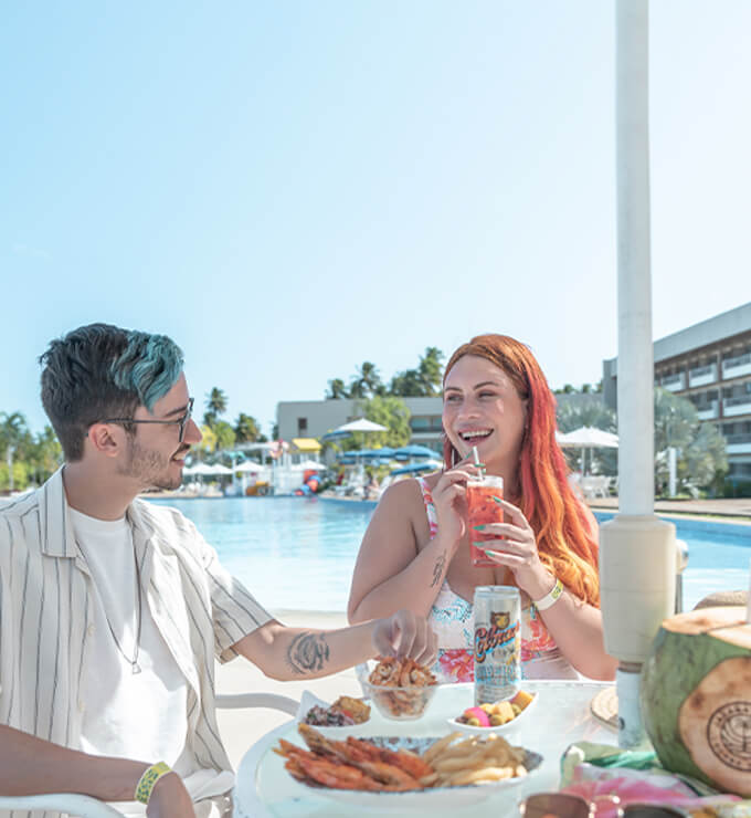 casal em um dos guarda-sóis na área comum da piscina, onde o homem esta bebendo uma cerveja colorado e a mulher um drink, na mesa também há petiscos para ambos.