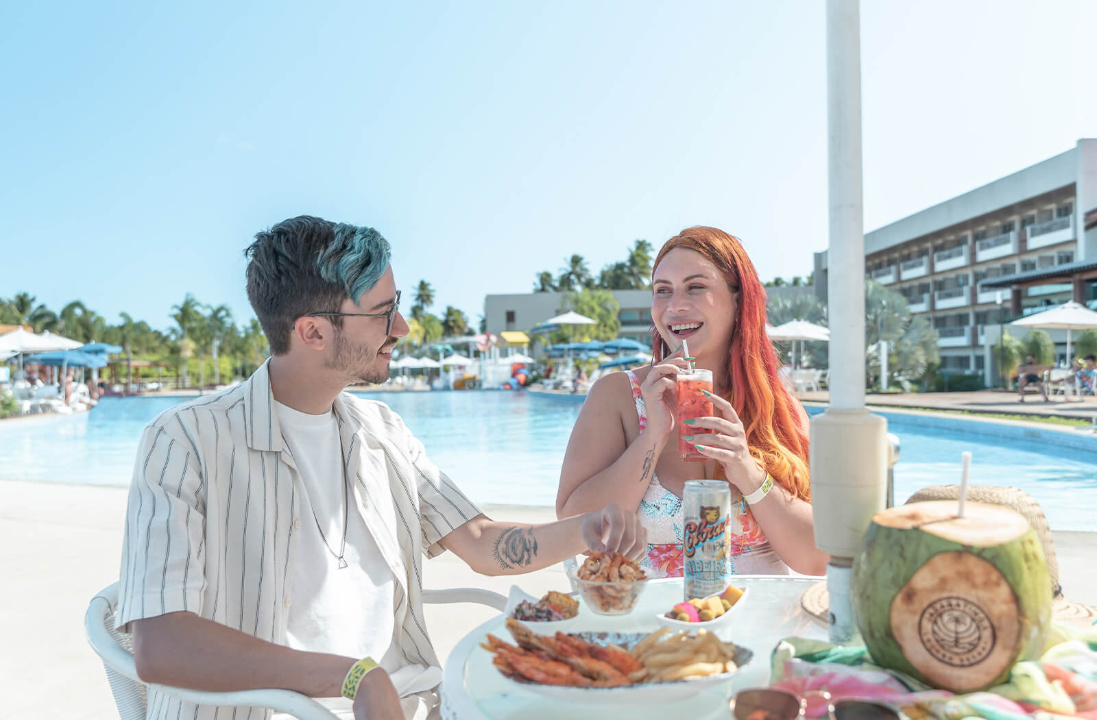 casal em um dos guarda-sóis na área comum da piscina, onde o homem esta bebendo uma cerveja colorado e a mulher um drink, na mesa também há petiscos para ambos.