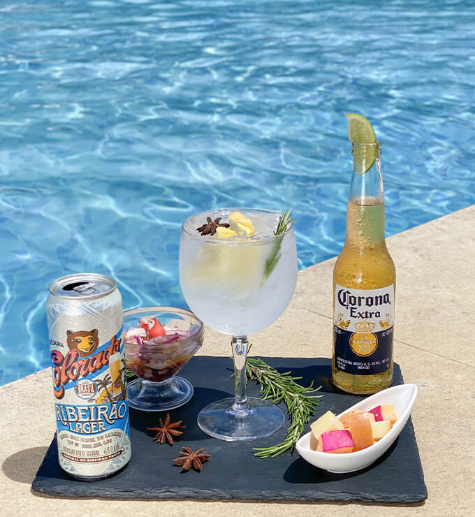 foto de uma bandeja servindo duas cervejas, uma corona e outra colorado na borda da piscina, também há um drink feito de vodka e alguns petiscos frios
