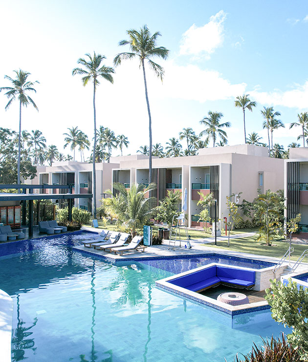 Vista da piscina do Garden Suíte, com o Garden e o Mangô Bar ao fundo. Com áreas de relaxamento, cercado por palmeiras e céu com poucas nuvens.