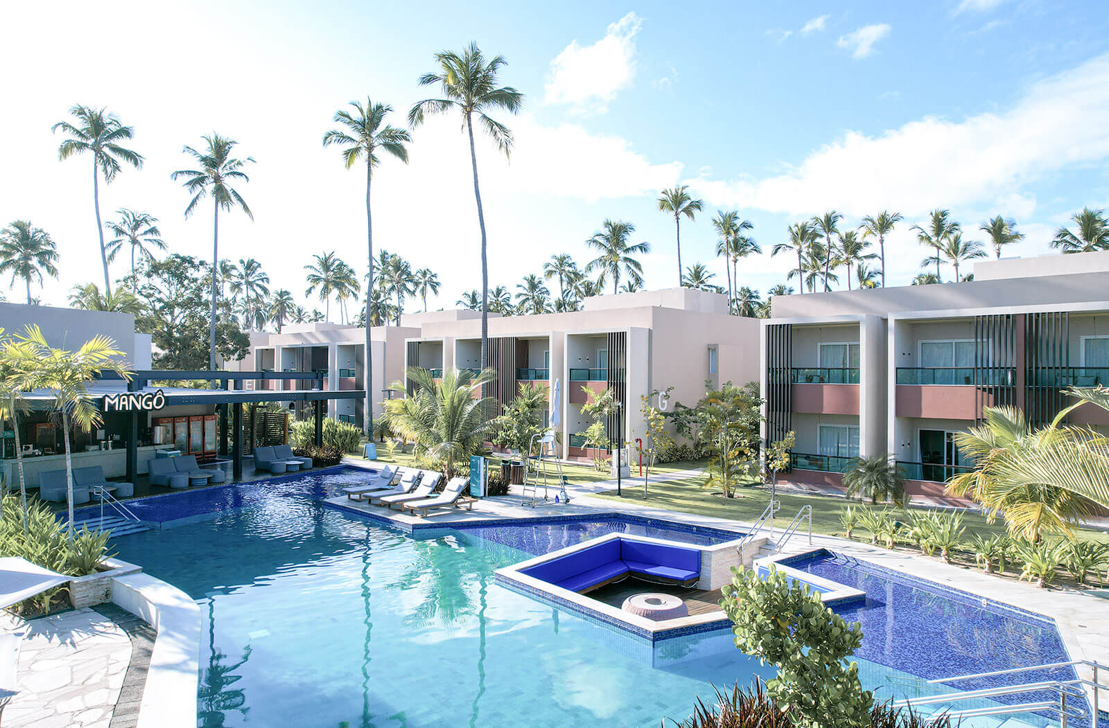 A imagem mostra uma vista aérea de parte do japaratinga resort. Há um conjunto de edifícios com arquitetura moderna, pintados em cores neutras com detalhes em terracota, alinhados em torno de uma área de piscina central. Espreguiçadeiras e guarda-sóis estão dispostos ao redor da piscina.