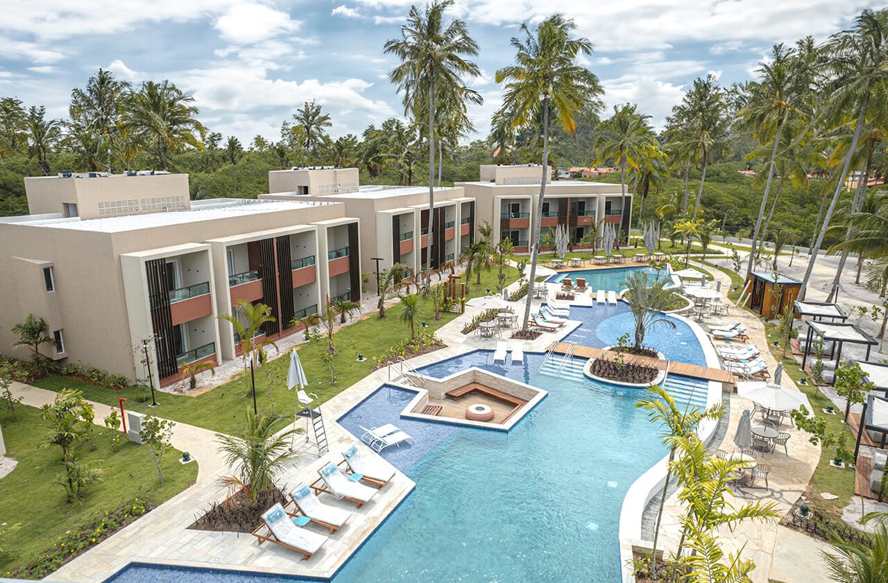 A imagem mostra uma vista aérea de parte do japaratinga resort. Há um conjunto de edifícios com arquitetura moderna, pintados em cores neutras com detalhes em terracota, alinhados em torno de uma área de piscina central. Espreguiçadeiras e guarda-sóis estão dispostos ao redor da piscina.