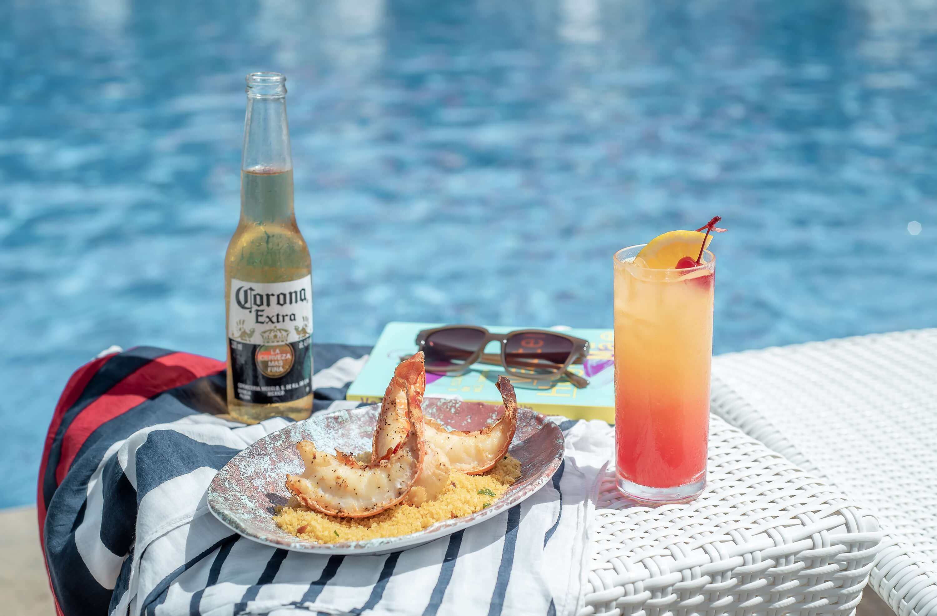 Pequena mesa na borda da piscina com cerveja Corona, porção de caudas de lagosta e drink Sex On The Beach. Canga, livro e óculos de sol ao redor.