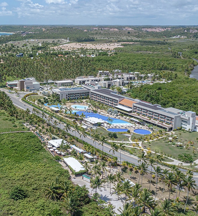 Vista aérea do Japaratinga Lounge Resort, com o complexo de piscinas e bangalôs entre a praia e um rio, com natureza integrada com todo o ambiente e céu com algumas nuvens.