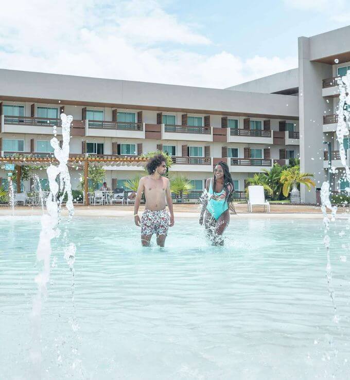 Homem e mulher caminham em piscina rasa, nas laterais chafarizes e ao fundo bloco de apartamentos