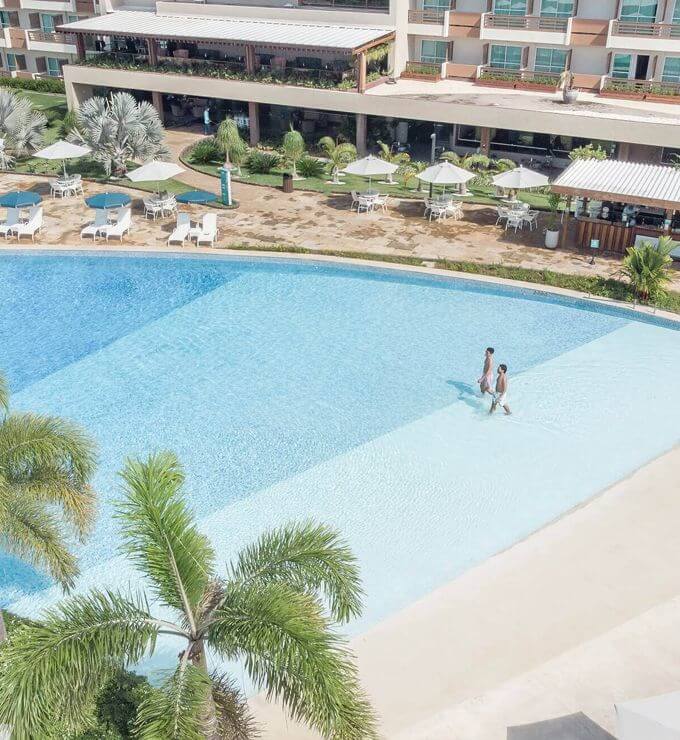 Vista aérea de dois homens entrando na piscina. Guarda-sóis, espreguiçadeiras e estrutura do resort ao fundo.